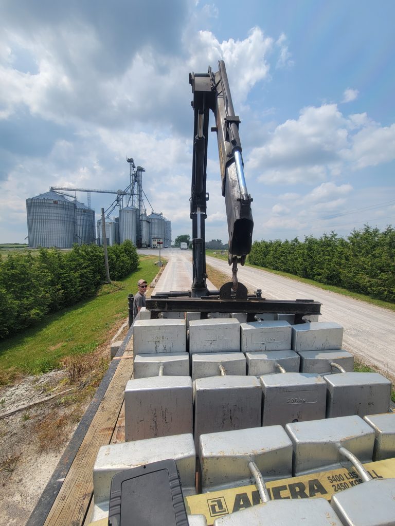 A crane is positioned over a flatbed trailer loaded with large metal weights, with grain silos in the background under a partly cloudy sky.